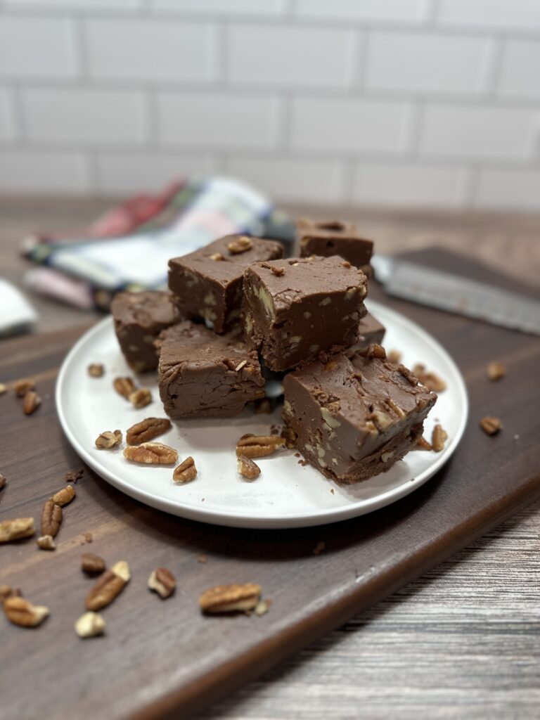 Pieces of pecan filled fudge on a white plate. The Plate is sitting on top of a wooden cutting board with pecans surrounding the plate.