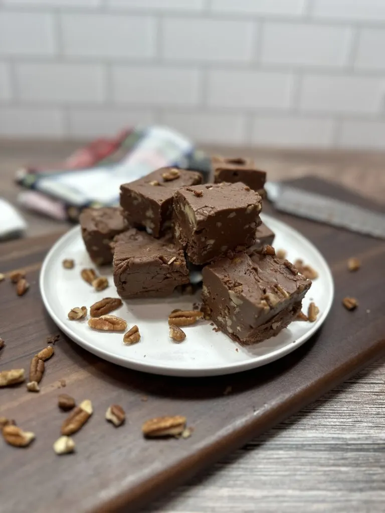 Pieces of pecan filled fudge on a white plate. The Plate is sitting on top of a wooden cutting board with pecans surrounding the plate.