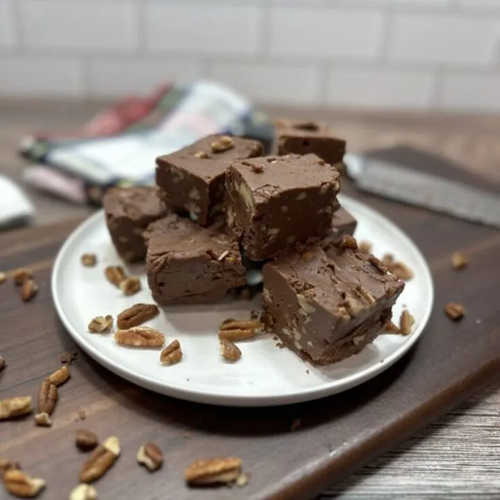 Pieces of fudge on a white plate. The Plate is sitting on top of a wooden cutting board with pecans surrounding the plate.