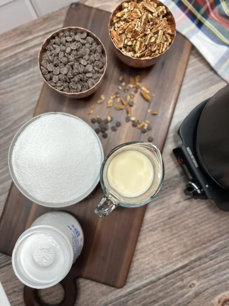 Pecans in a bowl, chocolate chips in a bowl, sugar in a bowl, evaporated milk in a cup, and fluffy marshmallow ingredients sitting on a cutting board.