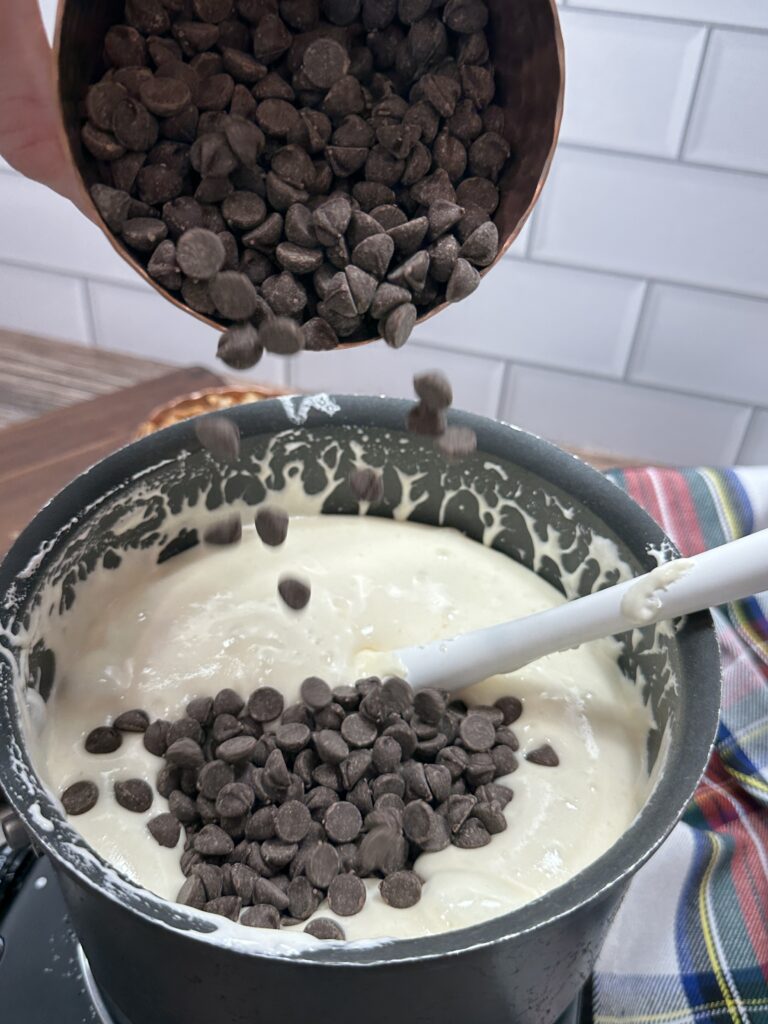 Cup containing chocolate chips being poured into the sauce pan with the stirred ingredients inside.