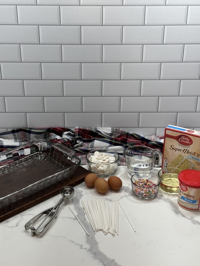 Image of all the simple ingredients of the recipe including candy melt, water, vegetable oil, vanilla cake mix, vanilla frosting, eggs, sprinkles, and lollipop sticks. A cookie cutter and a glass baking dish is also in the picture.