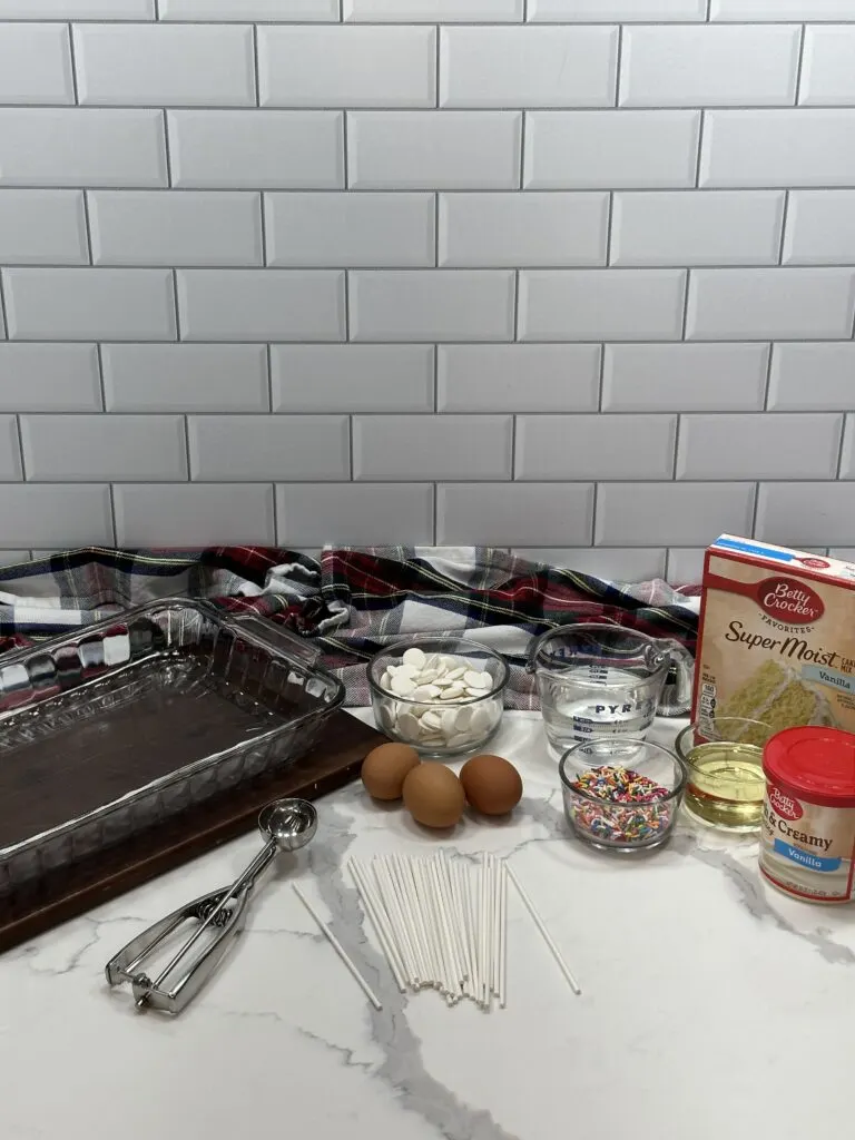 Image of all the simple ingredients of the recipe including candy melt, water, vegetable oil, vanilla cake mix, vanilla frosting, eggs, sprinkles, and lollipop sticks. A cookie cutter and a glass baking dish is also in the picture.