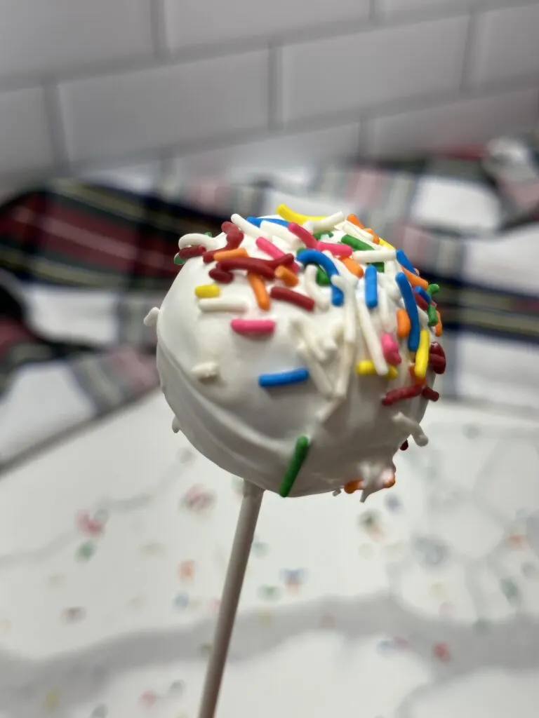 Image of 1 cake pop with vanilla frosting and rainbow sprinkles in the frame.