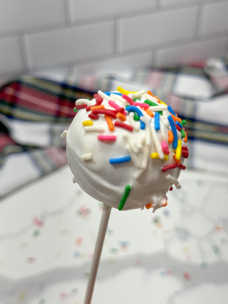 Image of 1 cake pop with vanilla frosting and rainbow sprinkles being held.