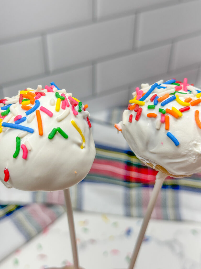 Image of 2 cake pops with vanilla frosting and rainbow sprinkles being held next to each other.